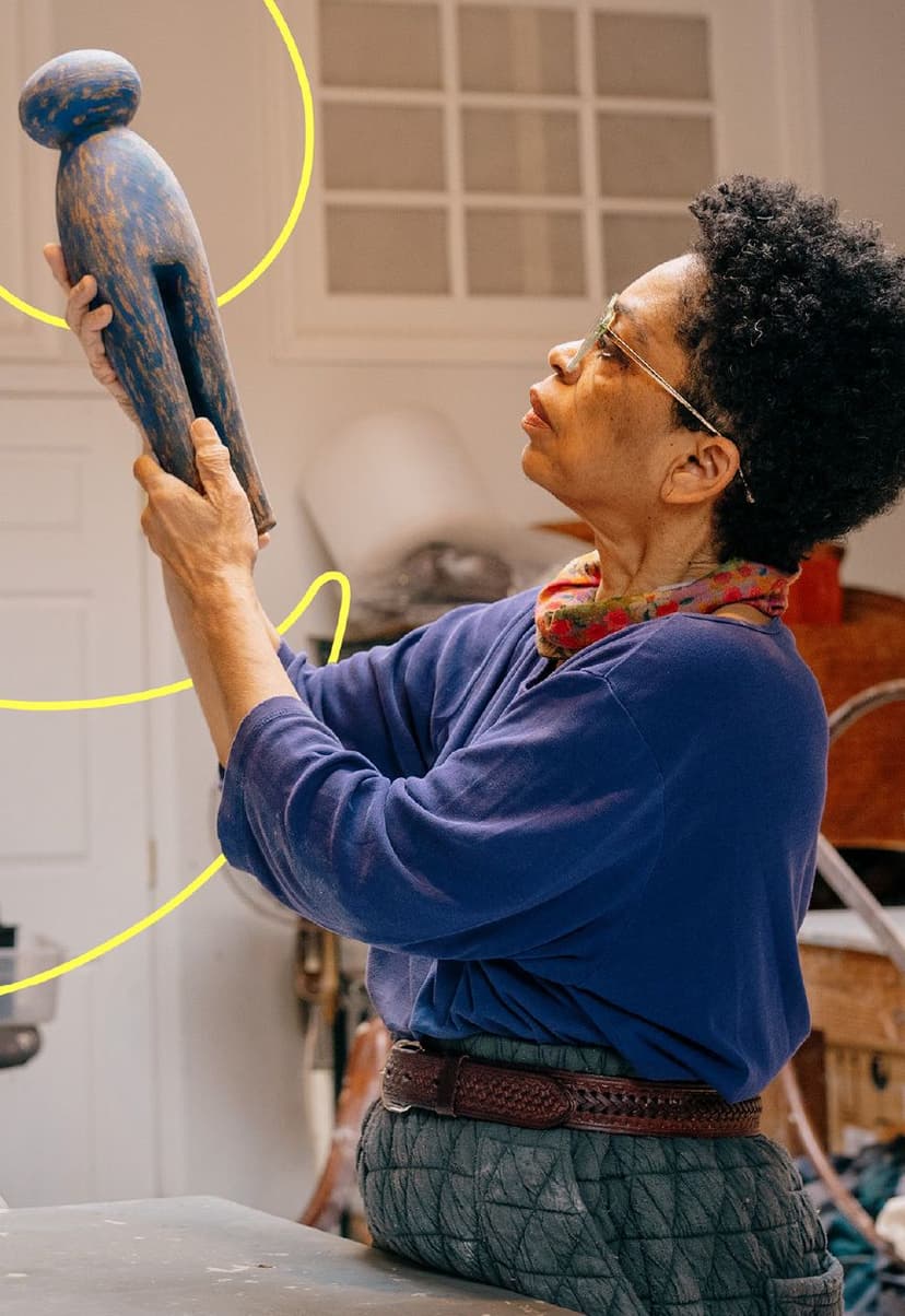 Person with short curly hair and glasses, wearing a blue long-sleeve shirt and a scarf, carefully examining a tall, blue wooden sculpture in an indoor setting.