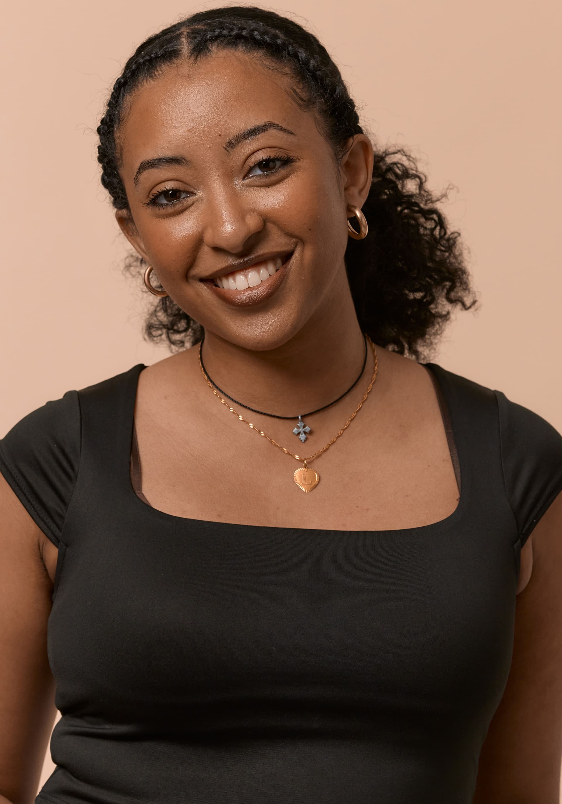 Young woman with curly hair tied back, wearing a black top and gold hoop earrings, smiling against a beige background. She has two necklaces, one with a circular pendant and the other with a cross.