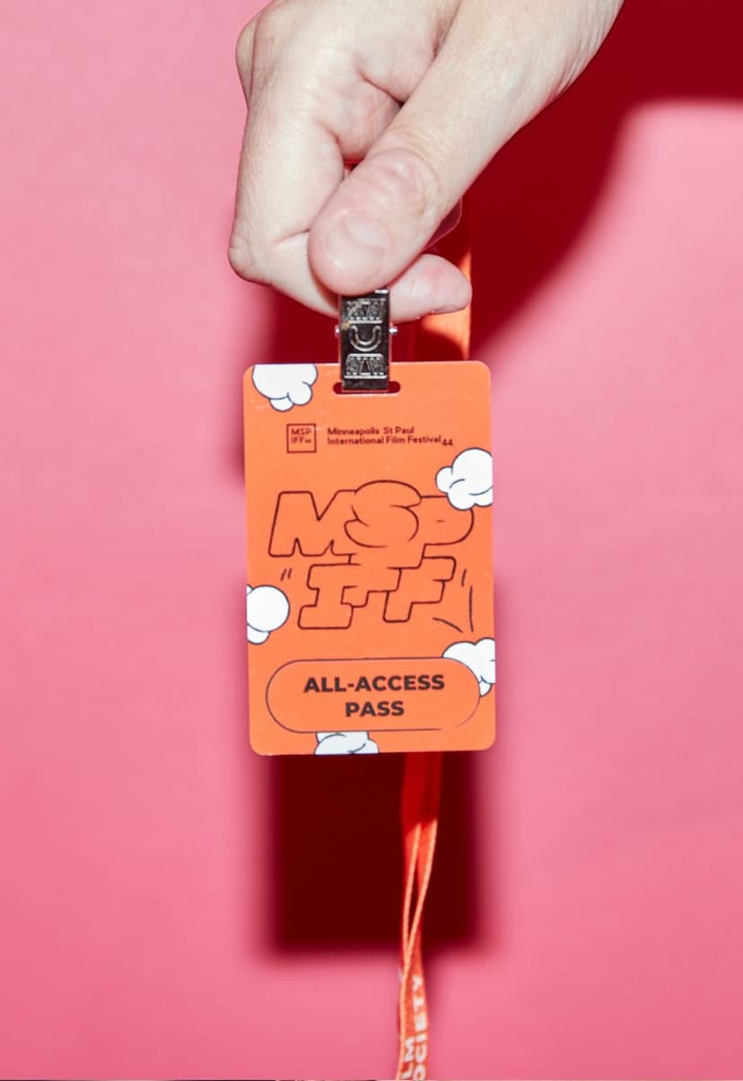 Hand holding an orange Minneapolis St Paul International Film Festival (MSP IFF) all-access pass with white cloud designs on a pink background.