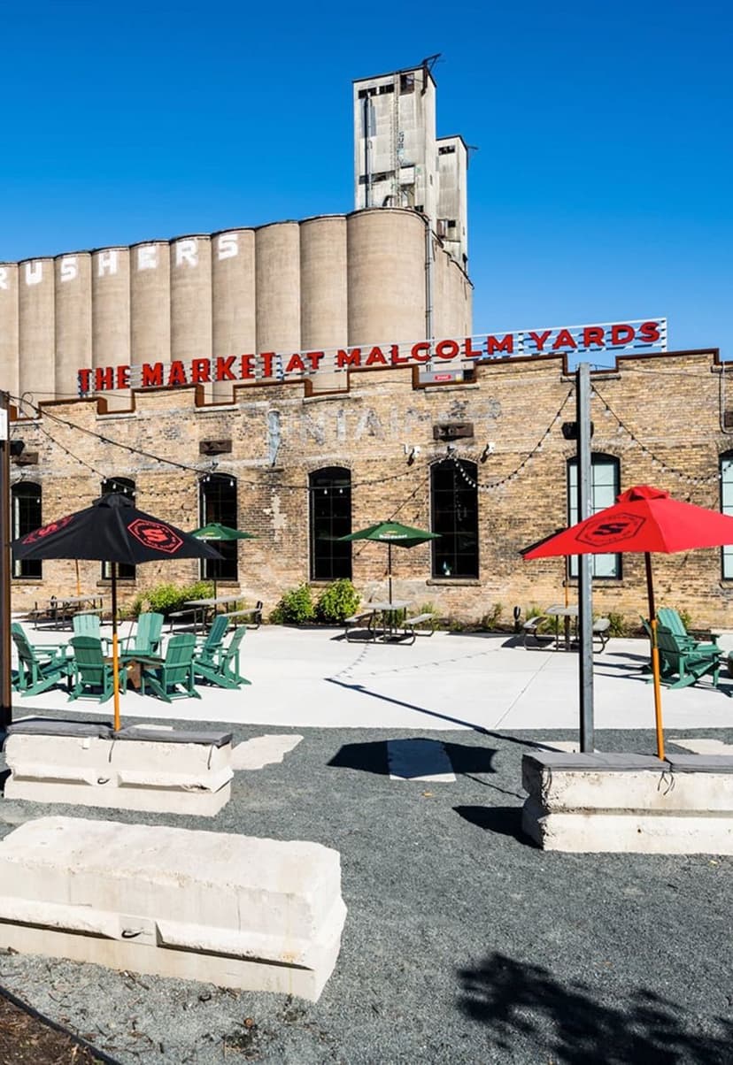 Outdoor seating area with green Adirondack chairs and picnic tables under black, green, and red umbrellas in front of a brick building with a sign reading "The Market at Malcolm Yards," with large industrial silos in the background under a clear blue sky.