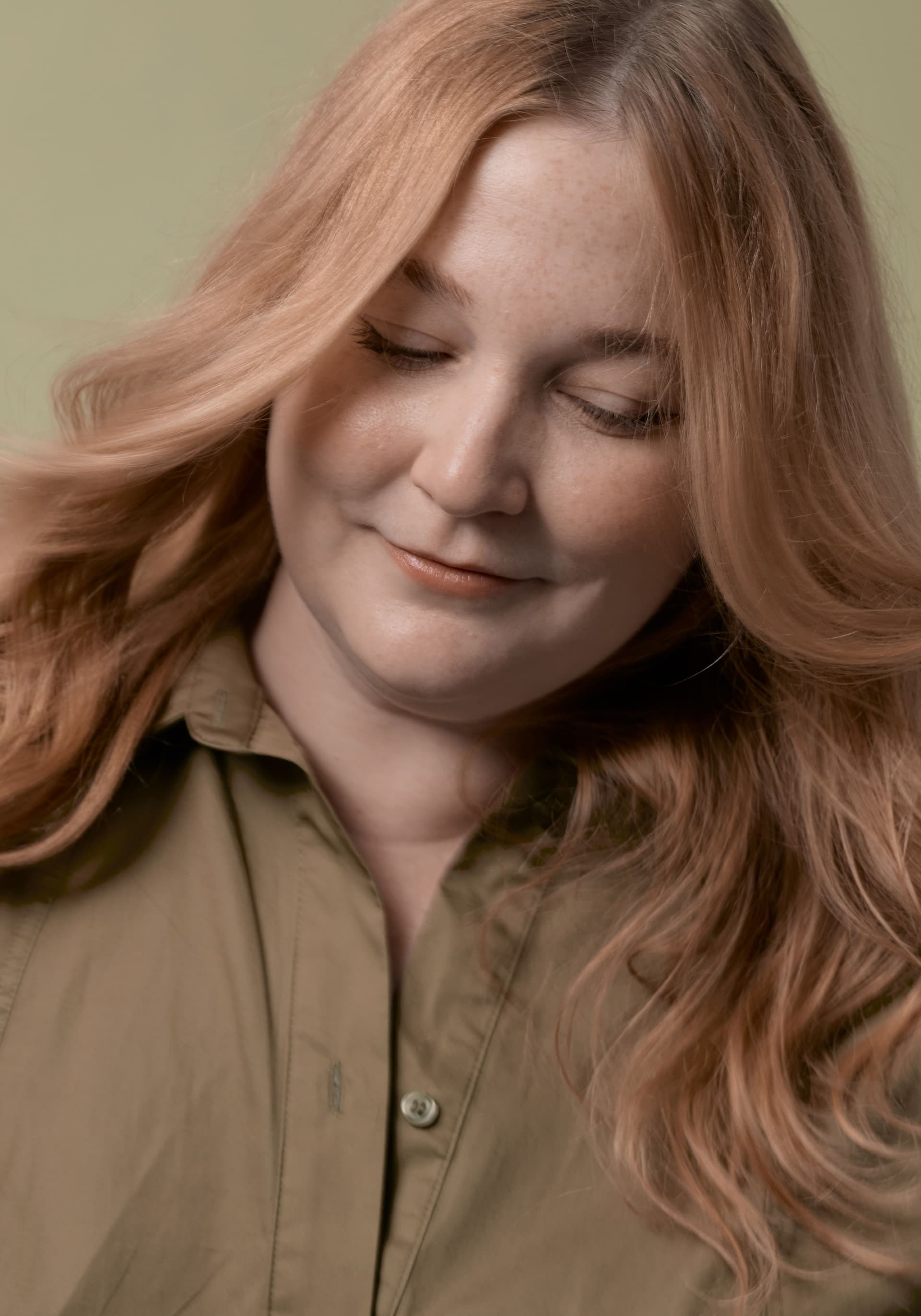 Close-up of a woman with long, wavy strawberry blonde hair, wearing a khaki green button-up shirt, looking down with a gentle smile against a light green background.