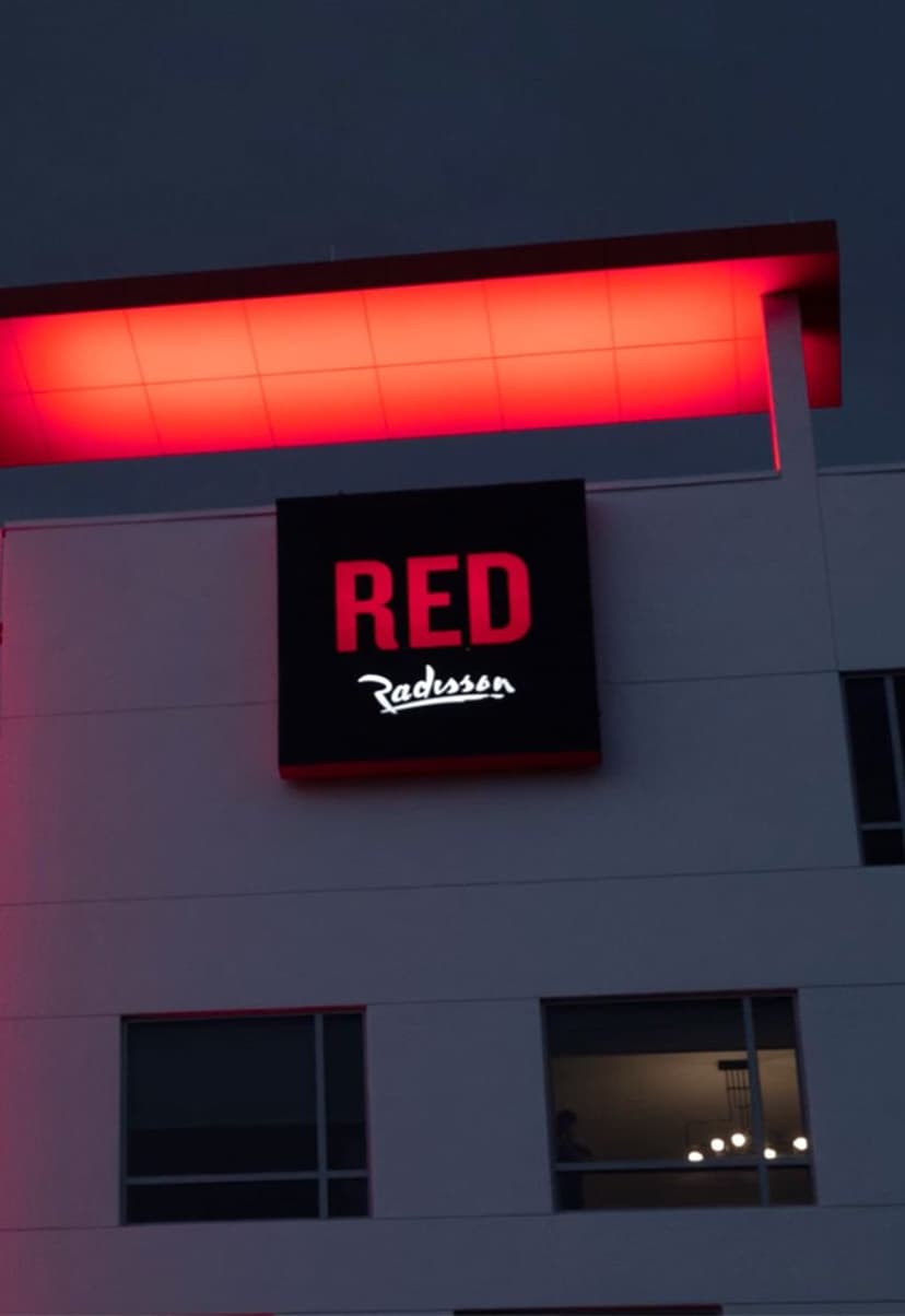 Illuminated Radisson RED hotel sign glowing in red on a modern white building at dusk, with a red-lit overhang and a lit chandelier visible through a window.