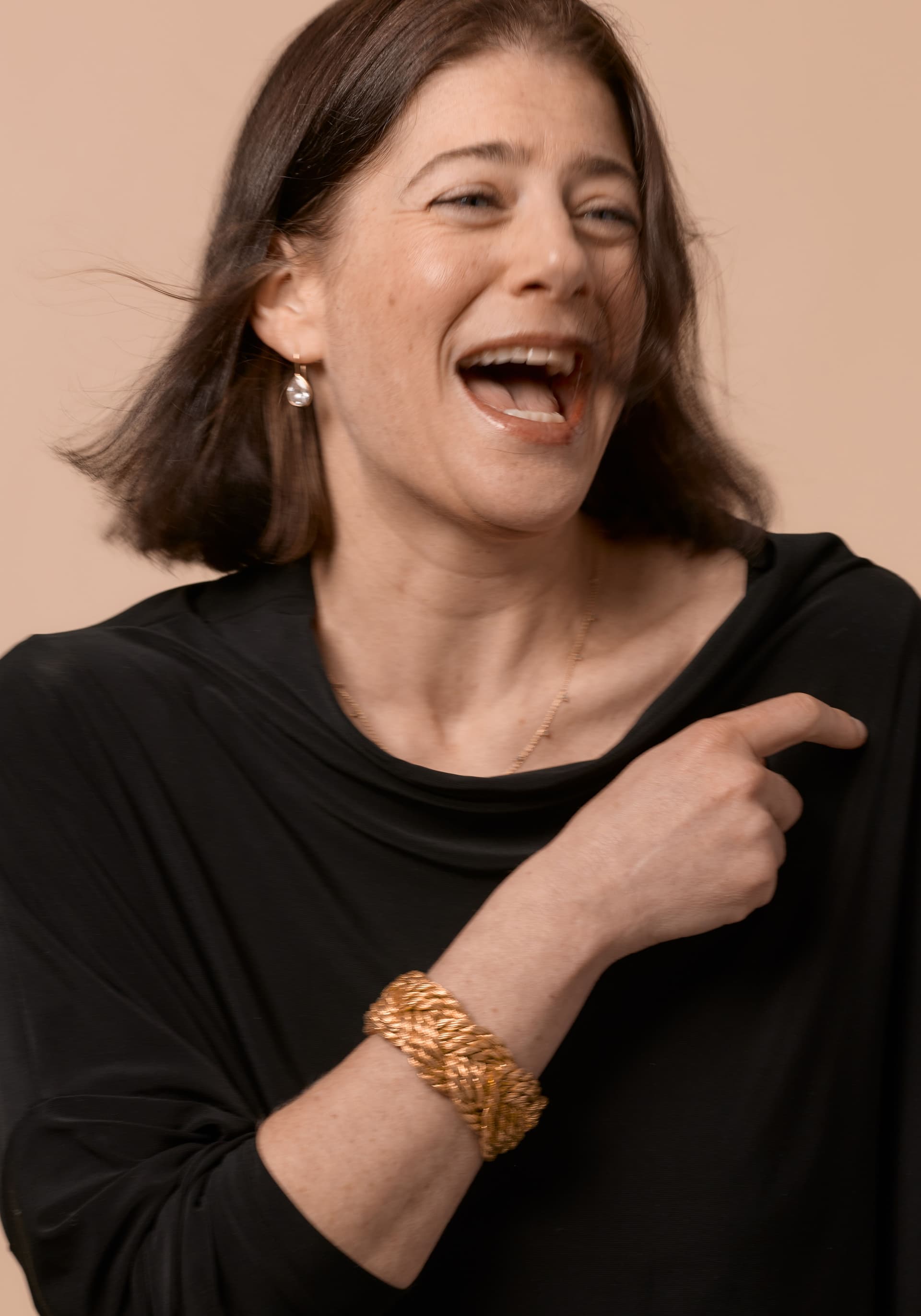 Woman with shoulder-length brown hair laughing, wearing a black top, a gold braided bracelet, and a pearl earring against a beige background.