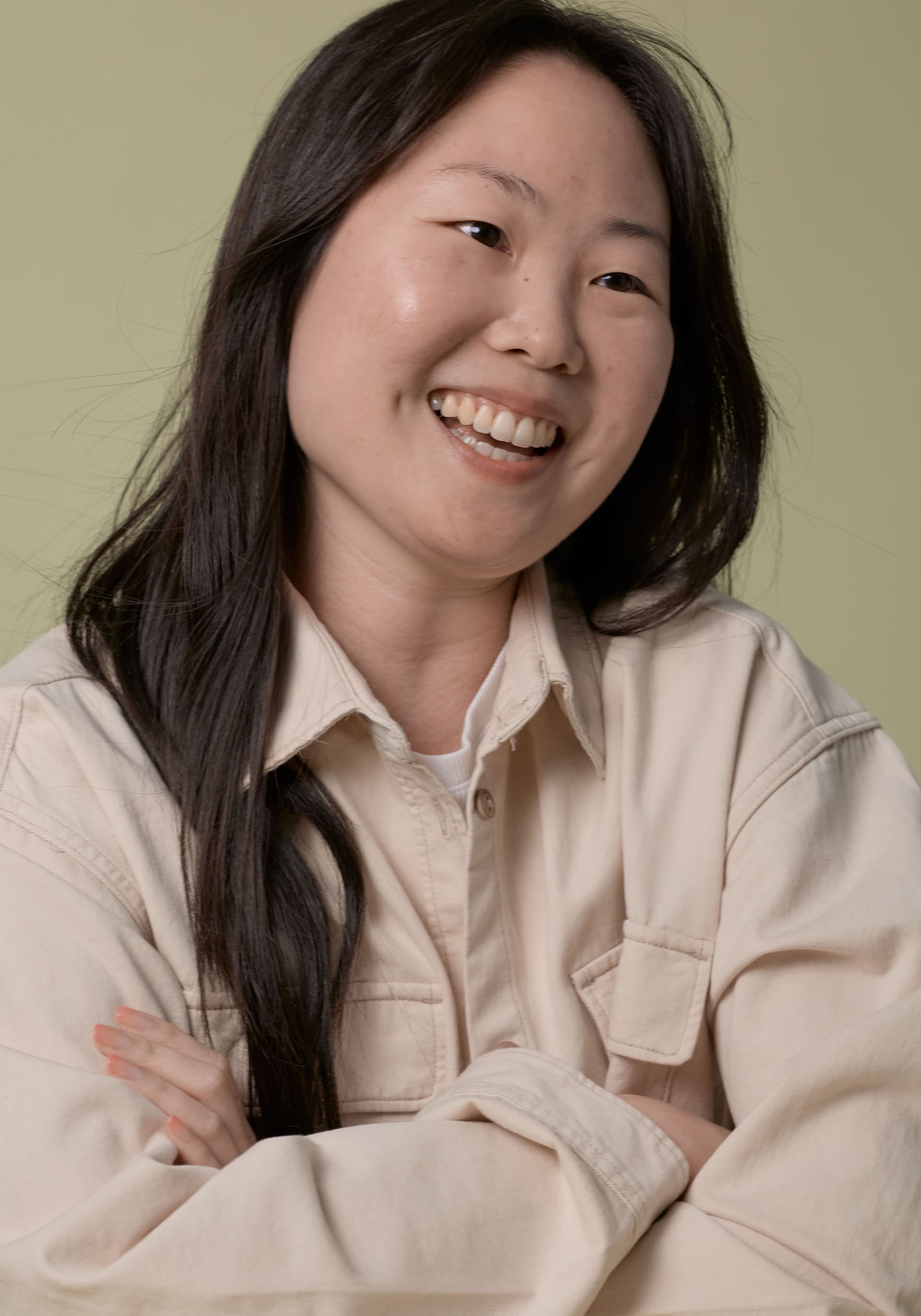 Portrait of a smiling woman with long dark hair, wearing a beige jacket over a white top, standing with arms crossed against a muted green background.
