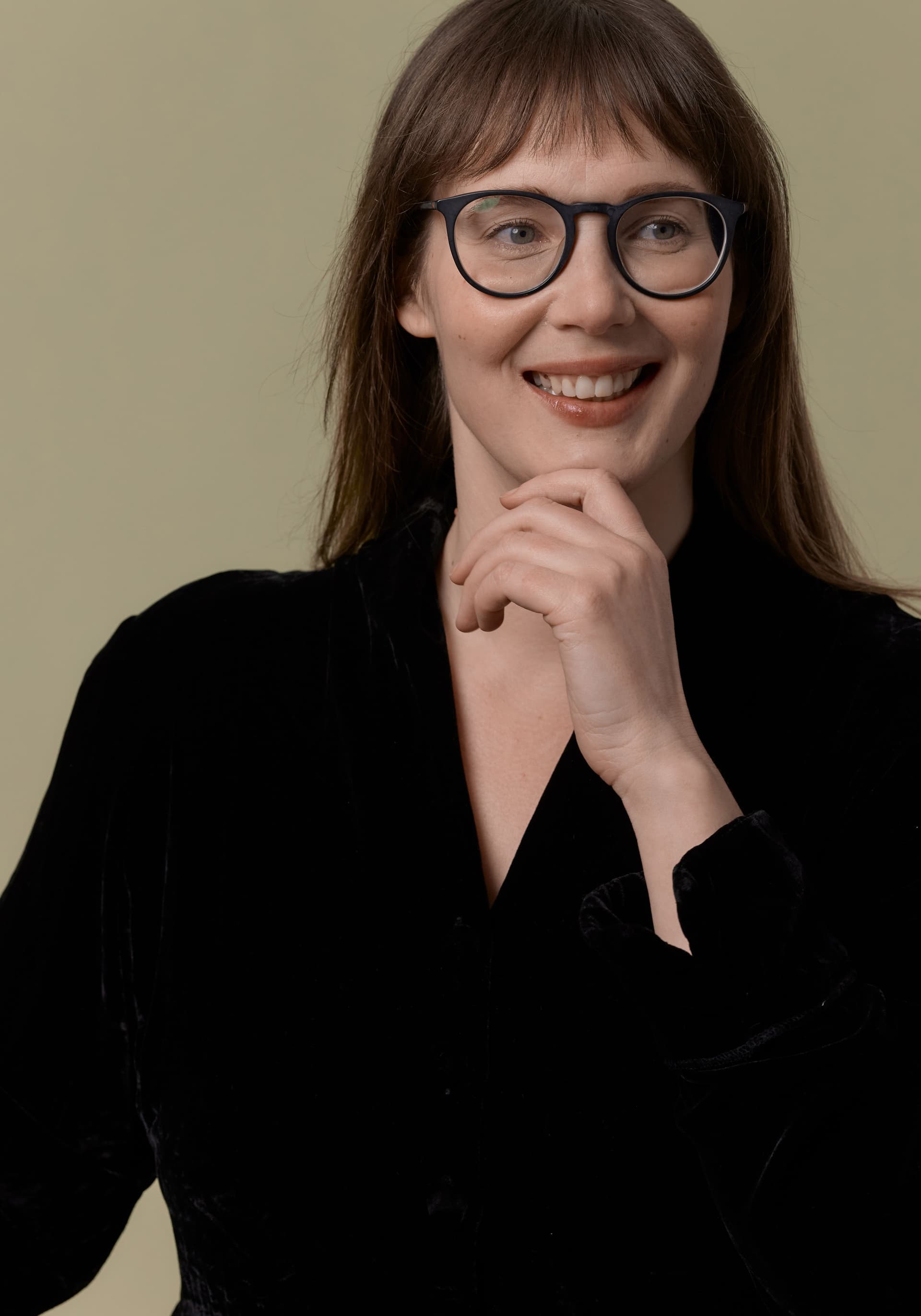 Portrait of a smiling woman wearing round black glasses and a black top, with her hand gently resting on her chin, against a plain olive-green background.