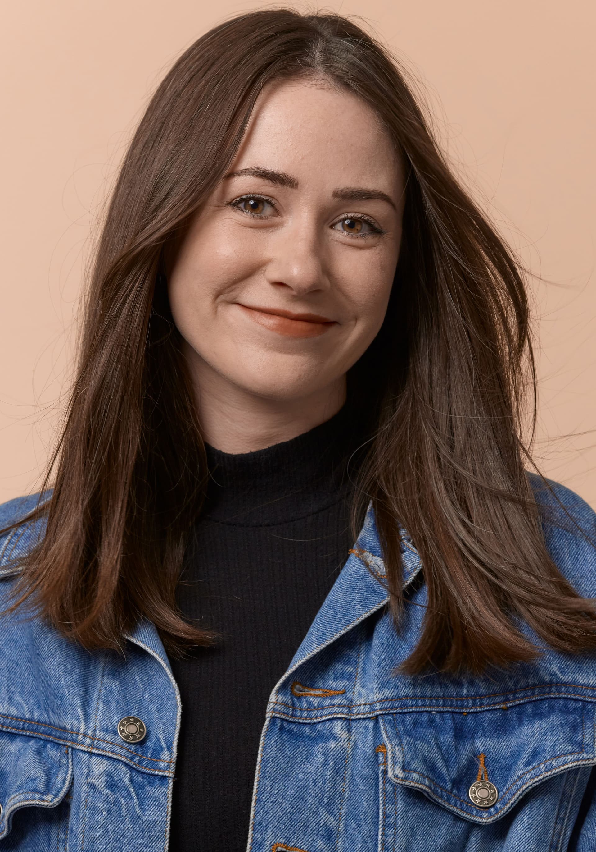 Portrait of a young woman with straight brunette hair, wearing a black turtleneck and a blue denim jacket, smiling softly against a light peach background.
